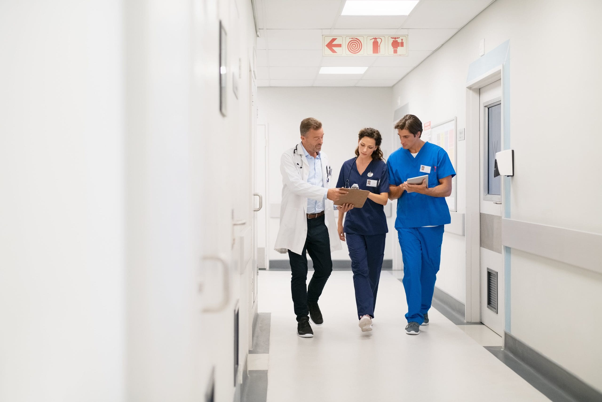 Three healthcare professionals walking down a hospital corridor, reviewing patient notes and discussing care.